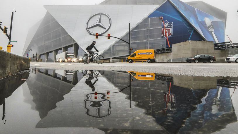 Work continued on Mercedes-Benz Stadium adhering Super Bowl graphics across the facade reflected in puddled water along Martin Luther King Jr. Drive in downtown Atlanta. A group of residents claims the stadium should be paying property taxes. JOHN SPINK/JSPINK@AJC.COM