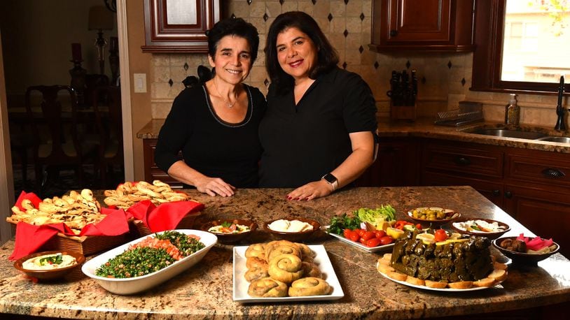 Sisters Hala Yassine (left) and Farrah Haidar pose with a few of the dishes they made: Akawi Twists (far left), Tabbouleh (center left), Maha’s Spiral Meat Pies (Sfeeha) (center) and Stuffed Grape Leaves (Warak Enab) (right). (Styling by Farrah Haidar and Hala Yassine / Chris Hunt for the AJC)