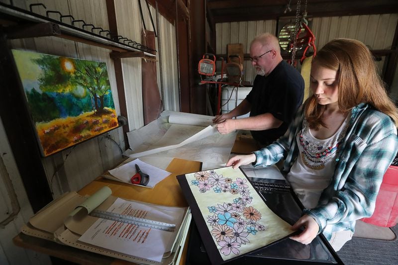 Mart Clamp and his daughter Lauren look over designs, including some of Lauren’s art work. The painting hanging on the wall of a tree in a forest was one Mart scooped out of the trash after Lauren had thrown it away. Curtis Compton/ccompton@ajc.com