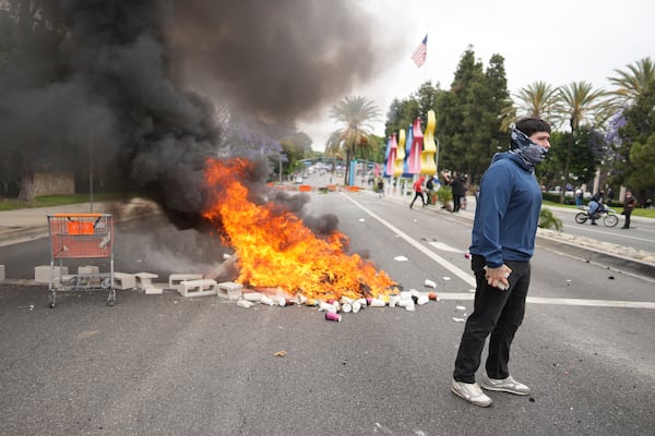 A fire burns as a protester stands across the way from Border Patrol personnel in riot gear and gas masks outside an industrial park in the city of Paramount, Calif., on Saturday, June 7, 2025 (AP Photo/Eric Thayer)