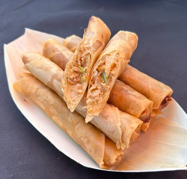 A paper boat dish holds a stack of thin fried spring rolls. Two are sliced and positioned so the viewer can see the inside of the rolls, which appears to be filled with shaved meat and green onions.