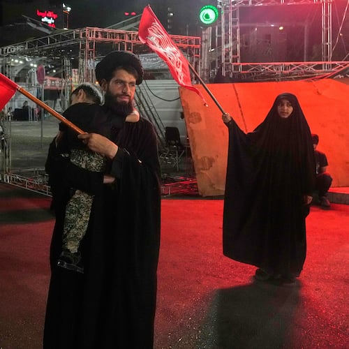 A cleric waves an Iranian flag while holding a child as a woman waves a religious flag in a pro-government gathering commemorating the late Iranian Supreme Leader Ayatollah Ali Khamenei at a square in Tehran, Saturday, July 11, 2026. (AP Photo/Vahid Salemi)