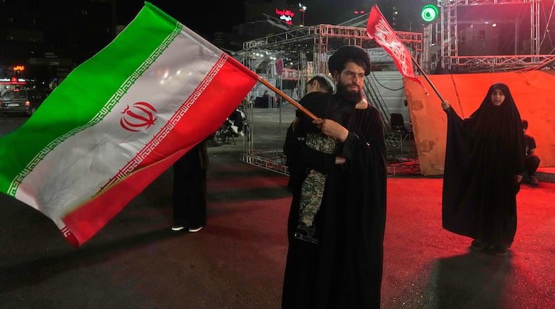 A cleric waves an Iranian flag while holding a child as a woman waves a religious flag in a pro-government gathering commemorating the late Iranian Supreme Leader Ayatollah Ali Khamenei at a square in Tehran, Saturday, July 11, 2026. (AP Photo/Vahid Salemi)