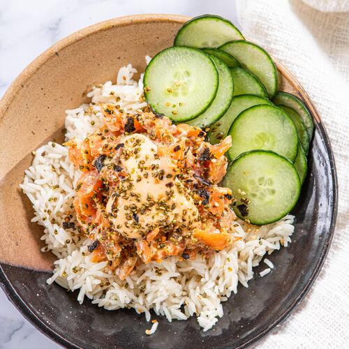A bowl of rice, salmon and cucumber slices, dressed with sriracha mayo and furikake, is placed on a marble countertop. A napkin is tucked under one side of the bowl, and a spoon rests on top of the napkin.