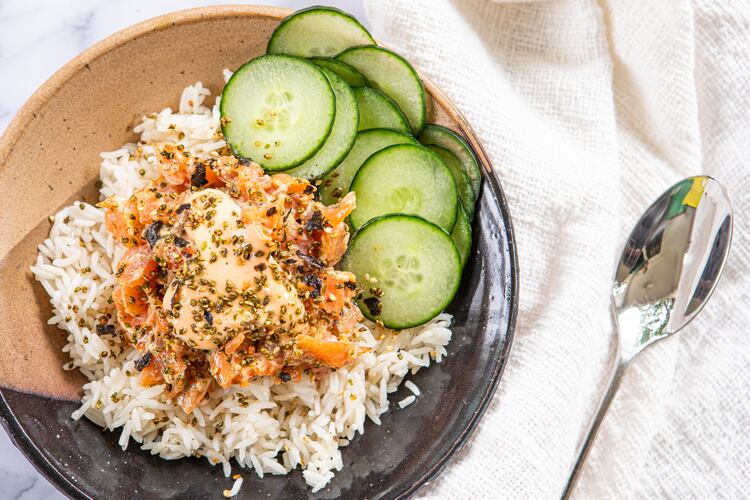 A bowl of rice, salmon and cucumber slices, dressed with sriracha mayo and furikake, is placed on a marble countertop. A napkin is tucked under one side of the bowl, and a spoon rests on top of the napkin.