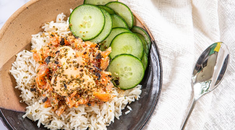 A bowl of rice, salmon and cucumber slices, dressed with sriracha mayo and furikake, is placed on a marble countertop. A napkin is tucked under one side of the bowl, and a spoon rests on top of the napkin.