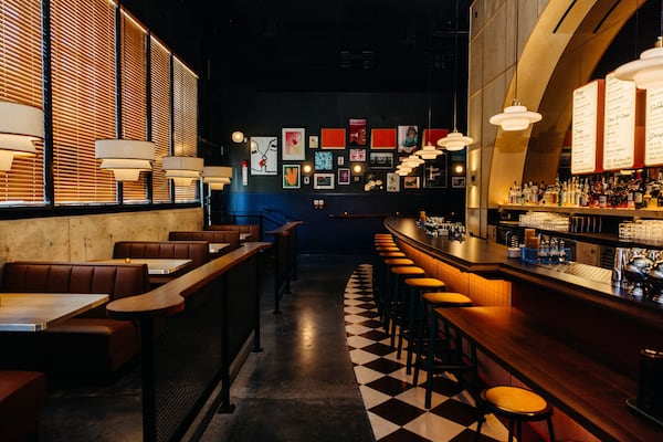 An empty barroom with retro styling, including a checkerboard tile floor under the bar, windows covered with blinds to the left and a gallery wall on the far wall.