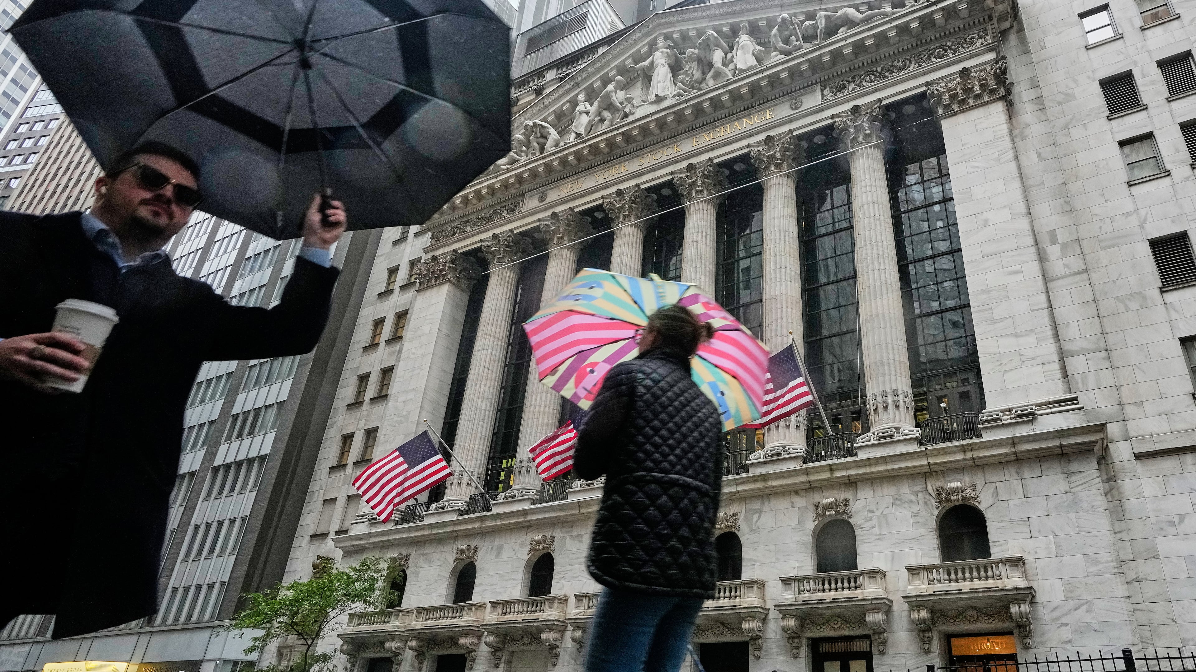 People with umbrellas pass the New York Stock Exchange, Monday, Oct. 13, 2025. (AP Photo/Richard Drew)