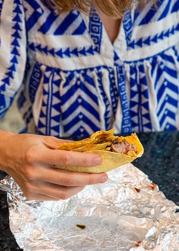A hand holds a steak taco with a bite taken out of the corn tortilla. The taco's foil wrapper is on the table below the taco.