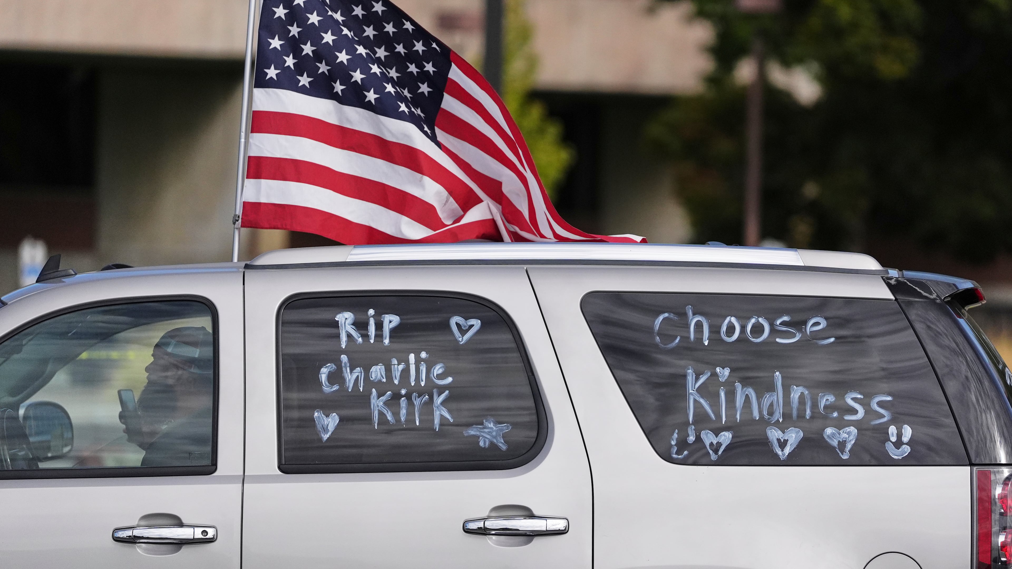 A vehicle marked with messages written on its windows in tribute to Turning Point USA CEO and co-founder Charlie Kirk and carrying an American flag, drive past at Utah Valley University, Saturday, Sept. 13, 2025, in Orem, Utah. (AP Photo/Lindsey Wasson)