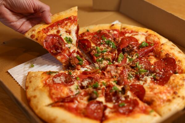 A hand is pulling a slice of pizza away from a full pie in a cardboard takeout box. The pizza is topped with pepperoni, sausage crumbles and chives.
