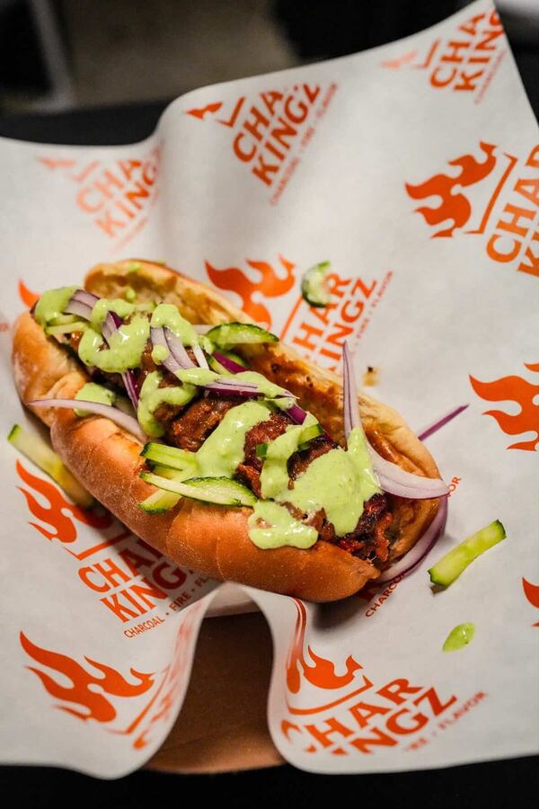 A beef kabob is presented like a hot dog and placed in a hot dog bun. The kabob is dressed with a sauce and slivers of red onion. The kabob and bun are resting on a paper food wrapper bearing the business name, Char Kingz.