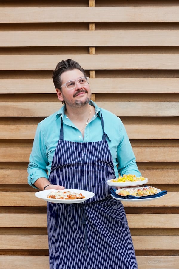 A bearded chef wearing an apron holds three plates of food, two on the right and one on the left.