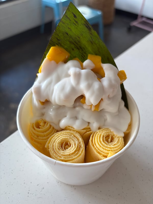 A cup of rolled ice cream topped with chunks of mango in a creamy sauce.
