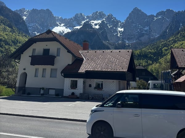 A van in Slovenia passes in front of a house with snow-capped mountains and clear blue skies in the background.