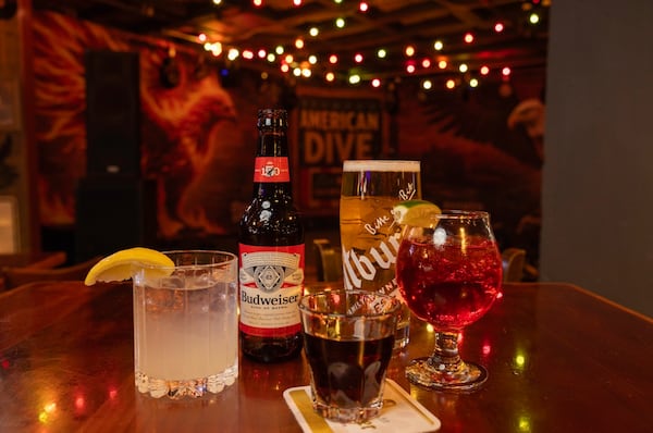 A wooden table framed by string lights holds three cocktails, a bottle of Budweiser and a glass of beer.
