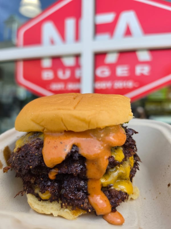 A large three-patty cheeseburger dripping with sauce on a plain bun on a disposable dish. The NFA burger logo appears behind the burger.