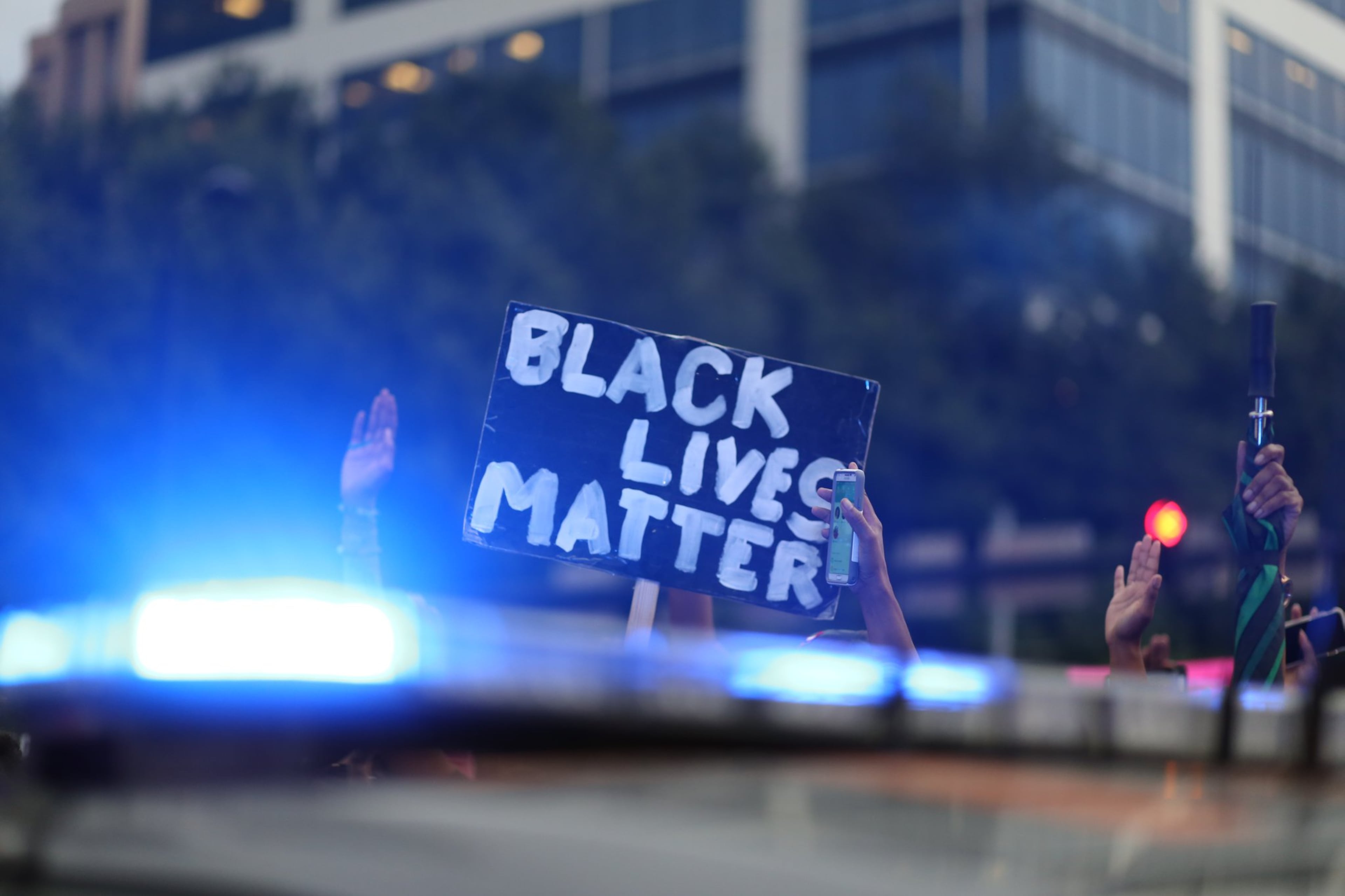 A police car's blue lights in the foreground with a Black Lives Matter sign held nearby in the summer of 2020. (Miguel Martinez/AJC)
