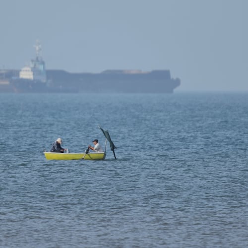 Two men sit in a small boat on the water as cargo ships are anchored in the background in the Strait of Hormuz off Bandar Abbas, Iran, Monday, May 4, 2026. (Amirhosein Khorgooi/ISNA via AP)
