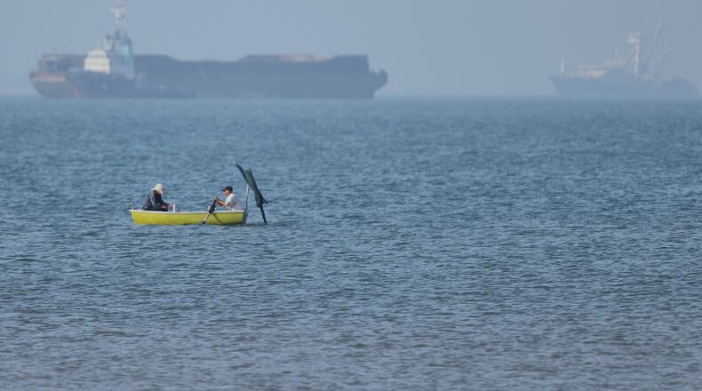 Two men sit in a small boat on the water as cargo ships are anchored in the background in the Strait of Hormuz off Bandar Abbas, Iran, Monday, May 4, 2026. (Amirhosein Khorgooi/ISNA via AP)