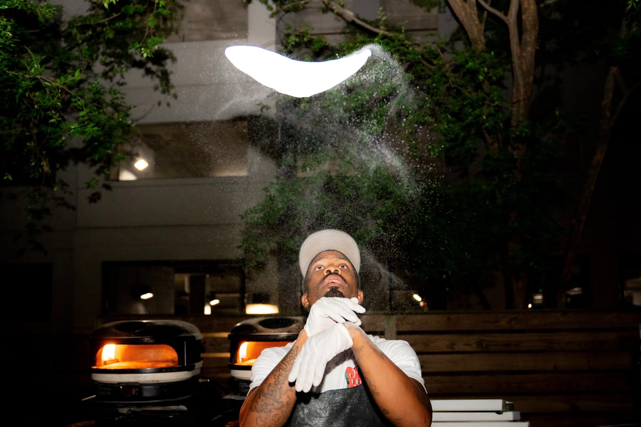 A man wearing rubber gloves and a hat tosses a pizza dough high above his head.