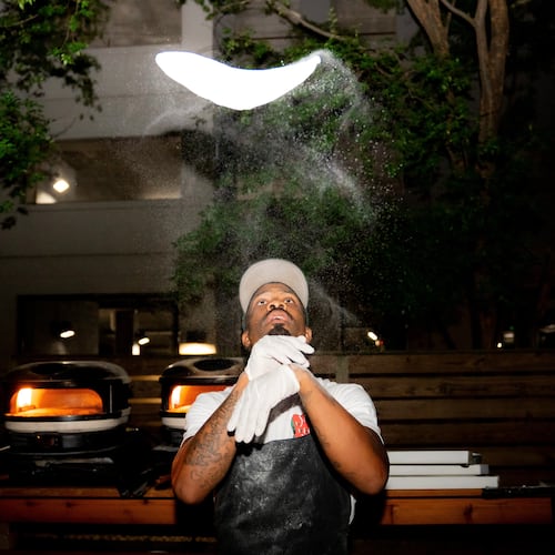 A man wearing rubber gloves and a hat tosses a pizza dough high above his head.