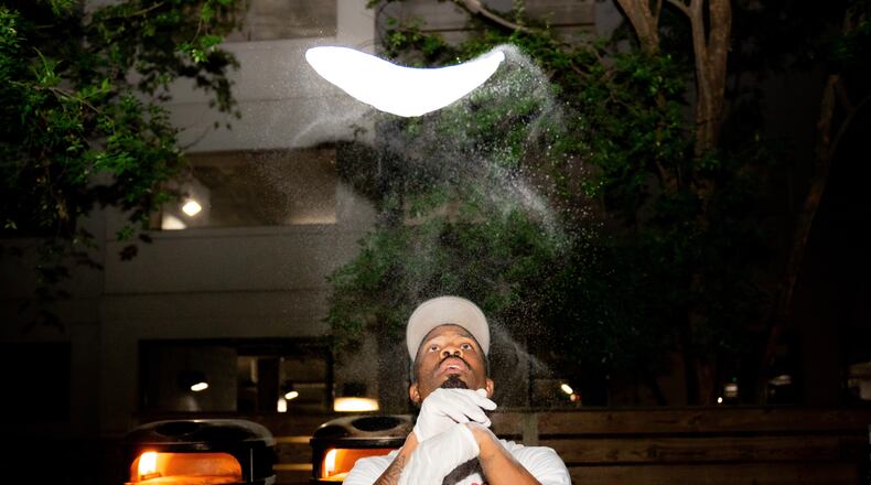A man wearing rubber gloves and a hat tosses a pizza dough high above his head.