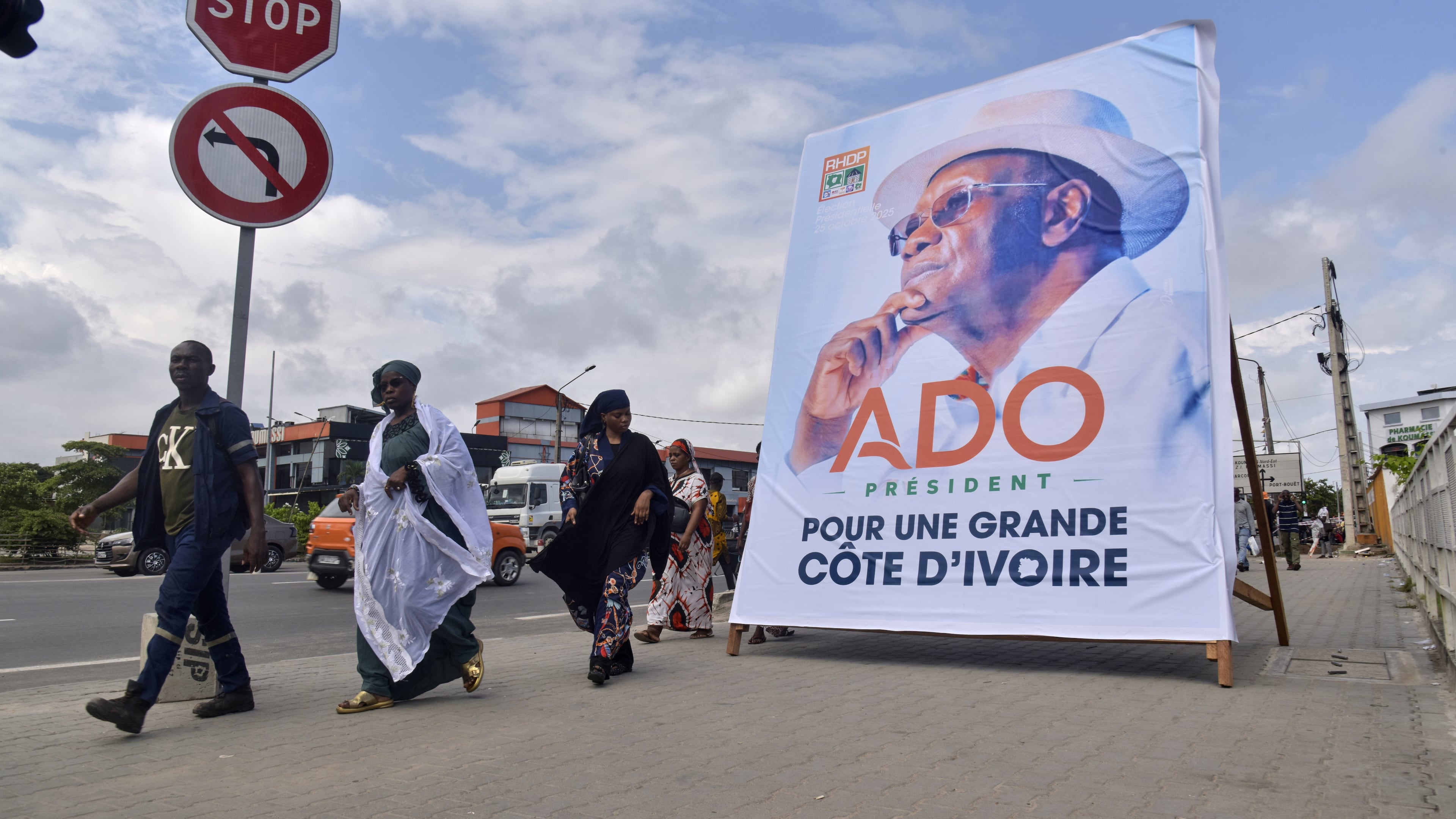 People walk past a campaign poster of President Alassane Ouattara in Abidjan, Ivory Coast, Friday, Oct. 10, 2025 (AP Photo/Diomande Ble Blonde)