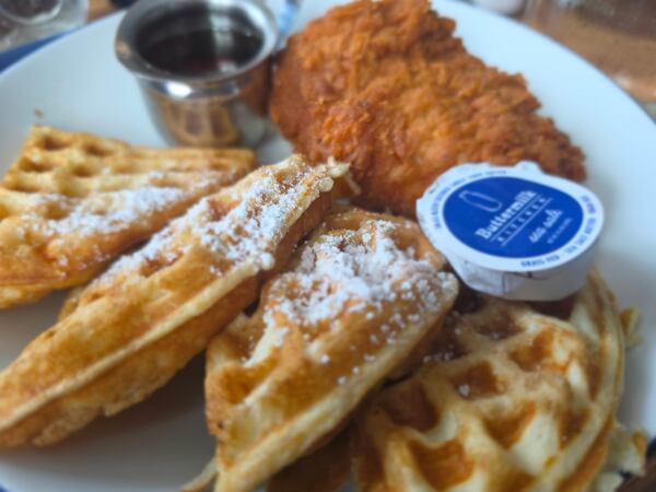 A closeup shot of a plate of chicken and waffles, with four waffle segments in front with a dash of powdered sugar and a large piece of fried chicken breast behind them, as well as a steel ramekin of syrup and an individually packaged pat of butter.