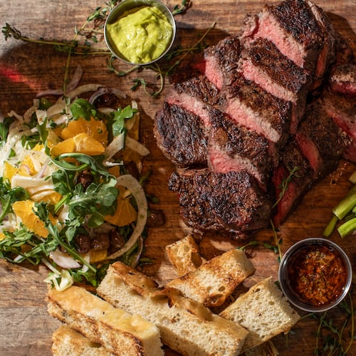 A sliced steak on a butcher board is surrounded with sides like pieces of grilled bread, potatoes, broccolini and a salad