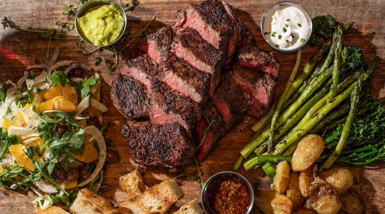 A sliced steak on a butcher board is surrounded with sides like pieces of grilled bread, potatoes, broccolini and a salad