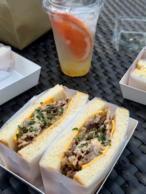 A paper-wrapped sandwich sliced in half to show generous amounts of braised beef, american cheese, and a sprinkle of green herbs. It is shown on a metal outside table, with a drink in the background.