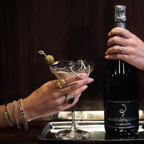 On the left, a woman's jeweled hand holds a martini garnished with olives and spoonful of caviar. On the right, another woman's jeweled hand holds a bottle of Champagne.