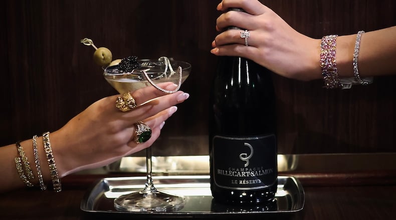 On the left, a woman's jeweled hand holds a martini garnished with olives and spoonful of caviar. On the right, another woman's jeweled hand holds a bottle of Champagne.