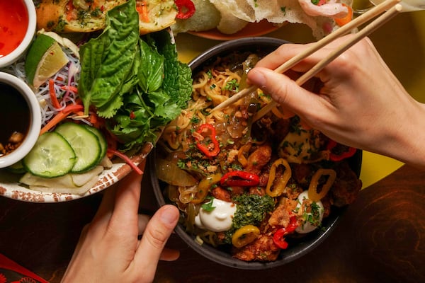 An overhead view of a bowl of ramen noodles topped with vegetables like sliced red and yellow peppers, with a bowl of vermicelli rice salad to the left topped with basil leaves, cucumbers and dressing. One hand holds the ramen bowl while another reaches in with chopsticks.