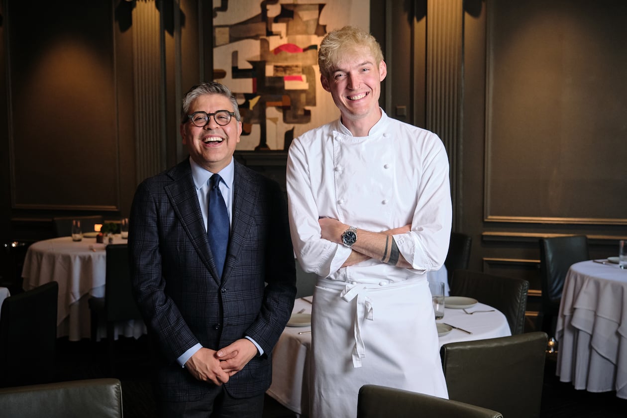 Two men stand in the dining room of a nice restaurant, the man of the left older and wearing a suit, the man on the right younger and wearing chef's whites.
