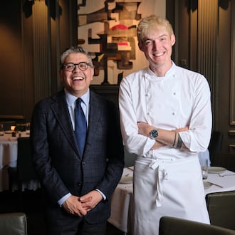 Two men stand in the dining room of a nice restaurant, the man of the left older and wearing a suit, the man on the right younger and wearing chef's whites.