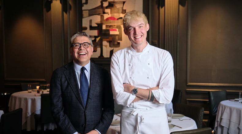 Two men stand in the dining room of a nice restaurant, the man of the left older and wearing a suit, the man on the right younger and wearing chef's whites.
