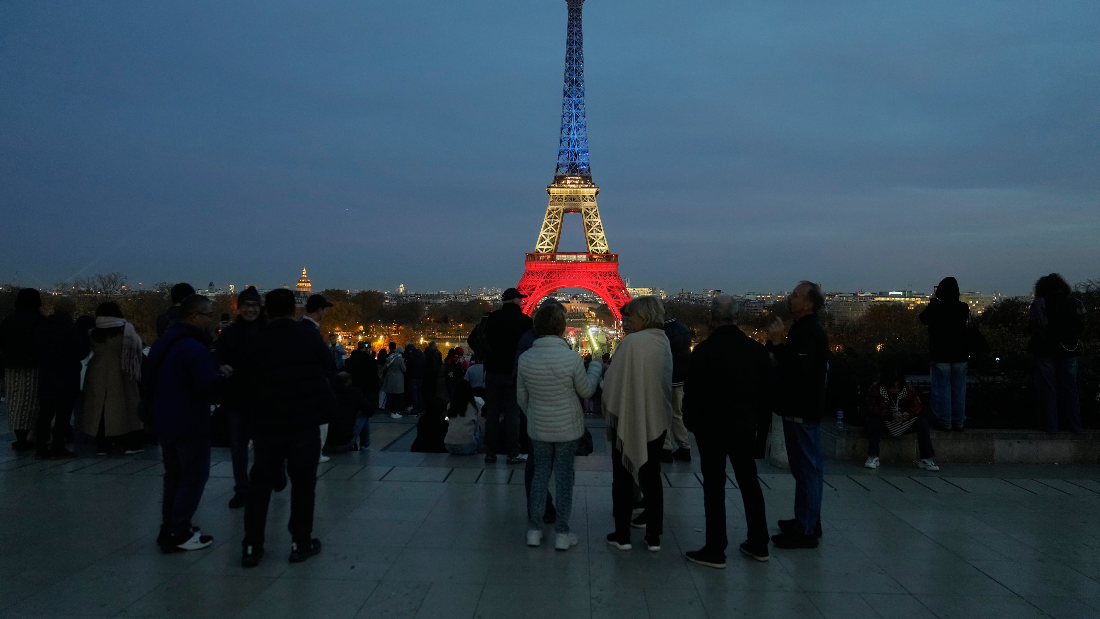 People gather at Trocadero Plaza as the Eiffel Tower is lit in the colors of the French national flag in Paris, Wednesday, Nov. 12, 2025, to honor the victims of the terror attacks at the Bataclan concert hall, cafes, and the national stadium 10 years ago. (AP Photo/Michel Euler)