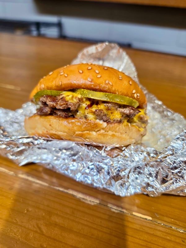 A 2-patty burger on top of its unwrapped aluminum foil. Melted cheese is settled into the crannies of the burgers. Two firm pickle slices peek out from the top of the sesame seed bun.