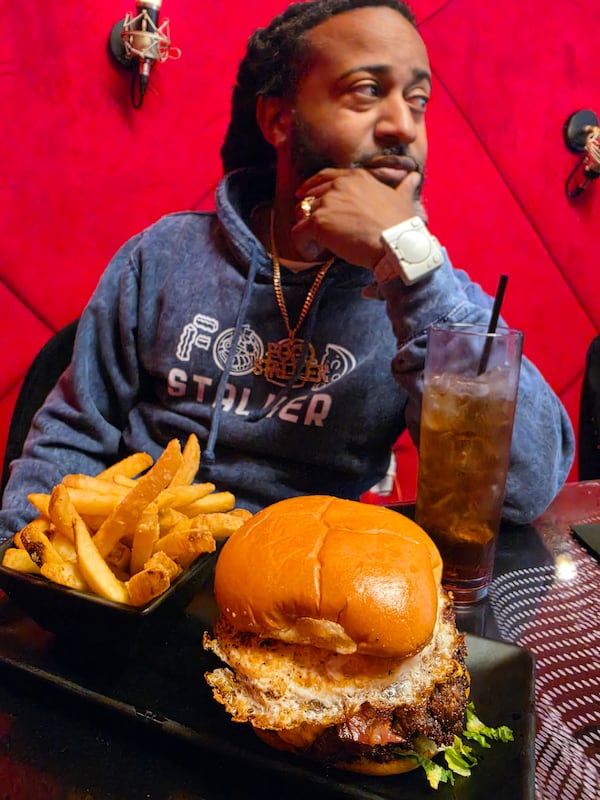 A large smash burger with a plain bun, fried egg and a little lettuce peeking out is accompanied by french fries. They are on a plate on a restaurant table. A man wearing a "Food Stalker" sweatshirt and stylish jewelry is posing in the background of the photo. He is resting his elbow on the table and his chin in his hand . A coke is next to his elbow.