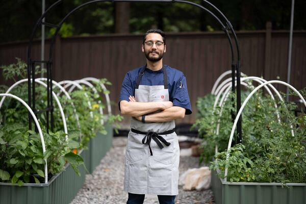 Fadi Alkaasamani is wearing a chef's apron and standing in between rows of tomato plants in a garden. His arms are folded across his body and he has a small smile on his face.