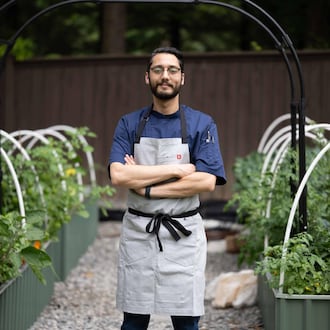 Fadi Alkaasamani is wearing a chef's apron and standing in between rows of tomato plants in a garden. His arms are folded across his body and he has a small smile on his face.