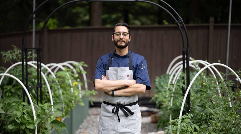 Fadi Alkaasamani is wearing a chef's apron and standing in between rows of tomato plants in a garden. His arms are folded across his body and he has a small smile on his face.