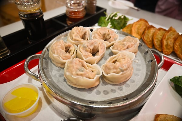 7 steamed dumplings on a stainless steel steamer, with fried dumplings, pickled radish and condiments in the background
