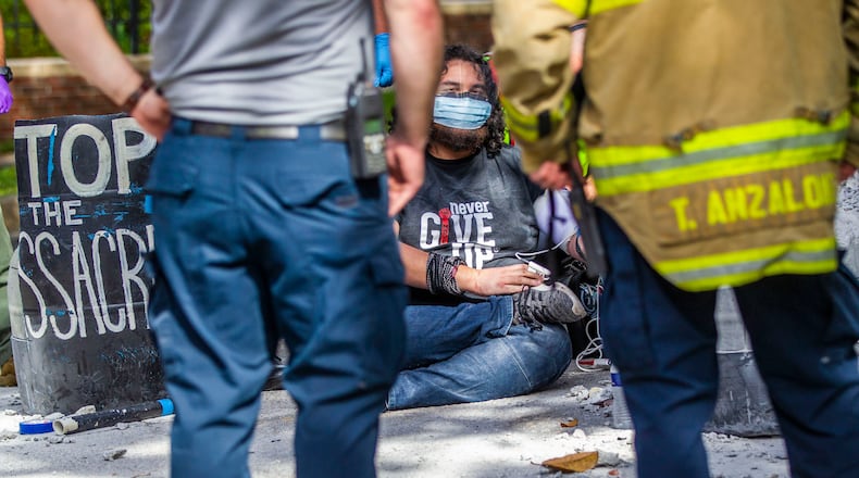 Protester Jordan Mazurek, 28, who cemented his hands in two 55-gallon plastic drums filled with concrete in the driveway of the Governor's Mansion, sits on the pavement while his vitals are checked by first responders, Friday, April 17, 2020, in Tallahasssee, Fla. Mazurek is protesting how Gov. Ron DeSantis is handling coronavirus in state prisons. (Alicia Devine/Tallahassee Democrat via AP)