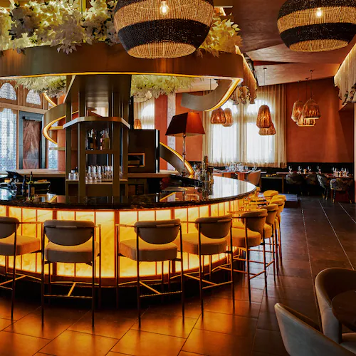 A warmly lit round bar in a hotel restaurant flanked by a wall of windows.