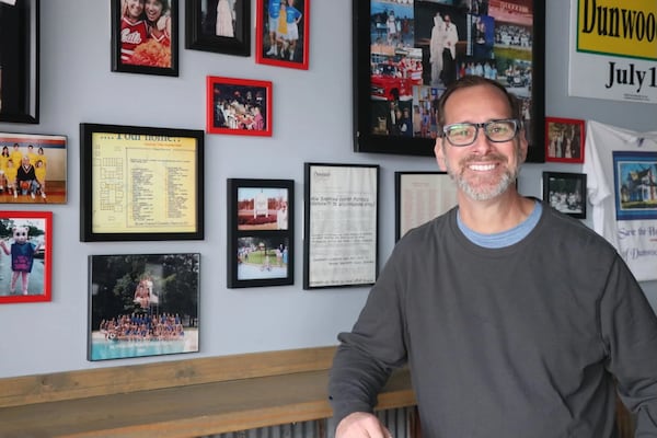 A man in a sweater stands smiling in front of a gallery wall of art.