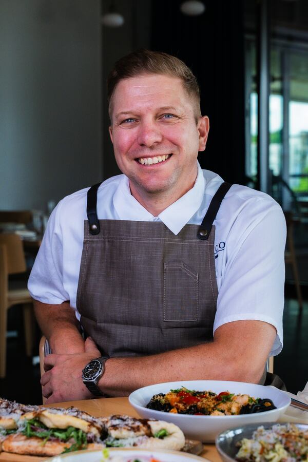 A male chef wearing an apron sits smiling at a table full of Italian food.