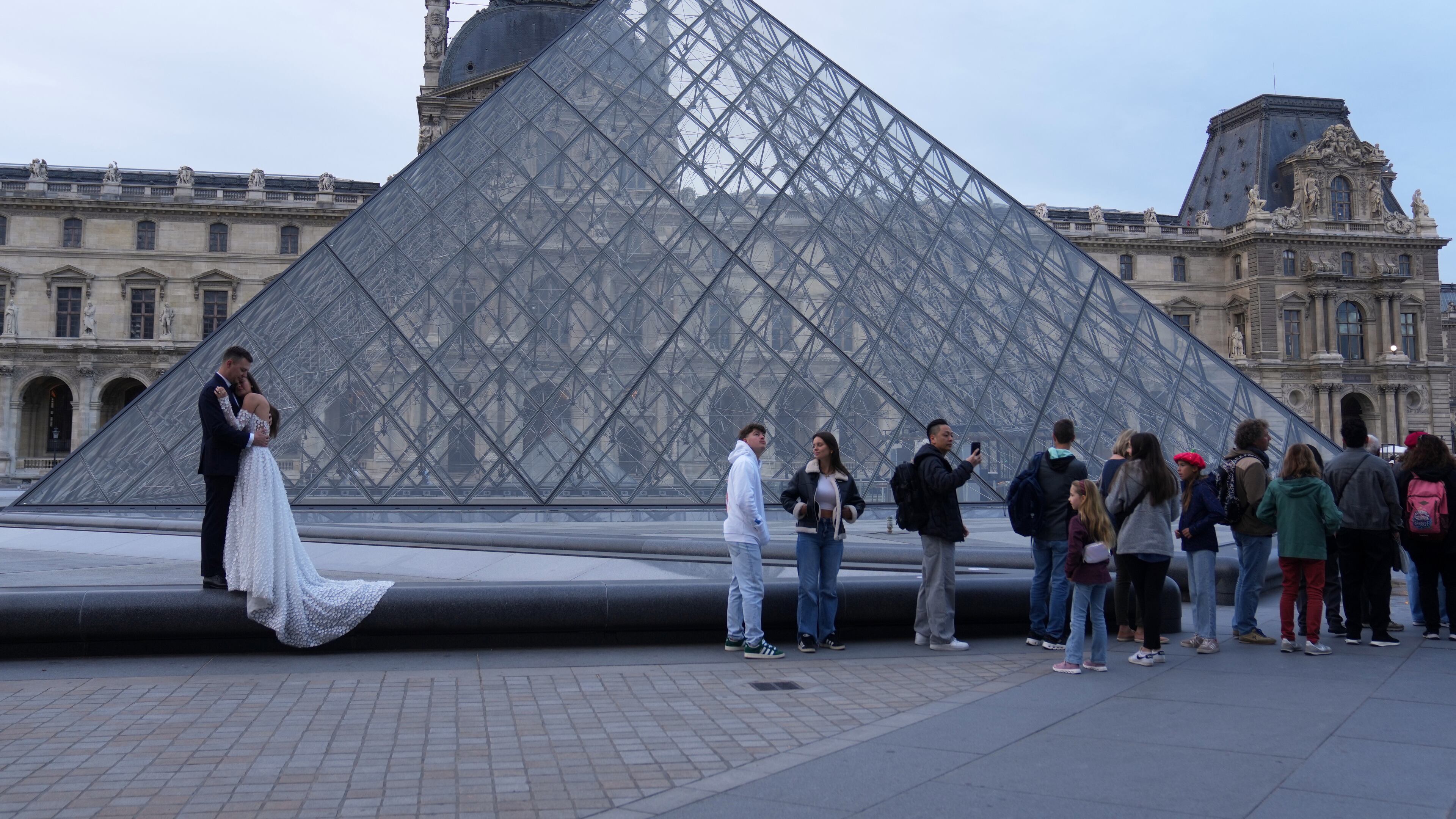 A wedding couple hugs as visitors queue to enter the Louvre museum three days after historic jewels were stolen in a daring daylight heist, Wednesday, Oct. 22, 2025 in Paris. (AP Photo/Thibault Camus)